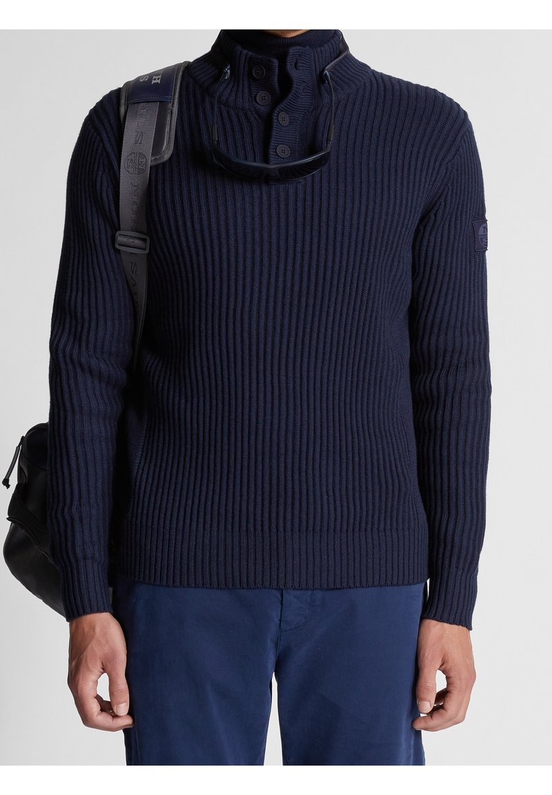Pull en tricot côtelé navy avec un col montant, une patte de boutonnage à trois boutons et un écusson logo sur l'épaule gauche. Associé avec un pantalon navy et un sac noir.