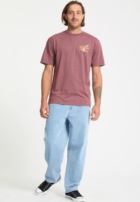 Bordeauxrotes Baumwoll-T-Shirt mit kurzen Ärmeln und einem Brustgrafik, kombiniert mit hellblauen Baggy-Jeans und schwarzen Sneakern.
