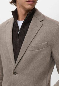 Blazer beige intrecciato con rever a punta, chiusura con un bottone e taschino sul lato sinistro del petto, indossato sopra un maglione a coste marrone scuro con colletto a zip.