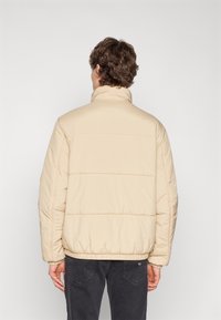 Beige puffer jacket with high collar, elastic cuffs, and hem. Features horizontal stitching and a smooth, slightly shiny texture.