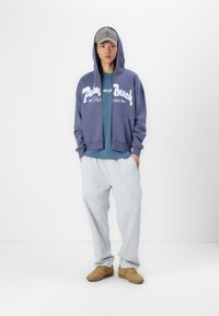 Hætte sweatshirt med lynlås i marineblå med hvid "Palm Beach" print, lagd over en teal skjorte. Lyseblå, løstsiddende bukser og brune sneakers.