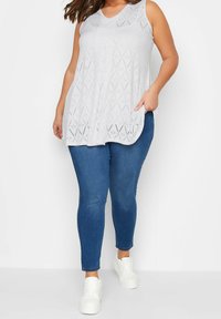 Kvinna som bär en vit ärmlös topp med broderi, blå smal jeans och vita sneakers, stående mot en enkel vit bakgrund.