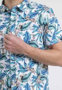 Chemise à manches courtes avec un fond blanc, ornée d'un motif de feuillage tropical coloré en teal et nuances de marron, avec un design à boutons.