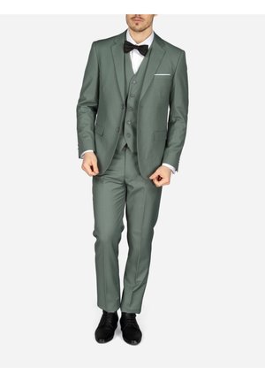 Man die een groen driedelig pak draagt met een zwarte strik, wit overhemd en zwarte nette schoenen, staand tegen een effen achtergrond.