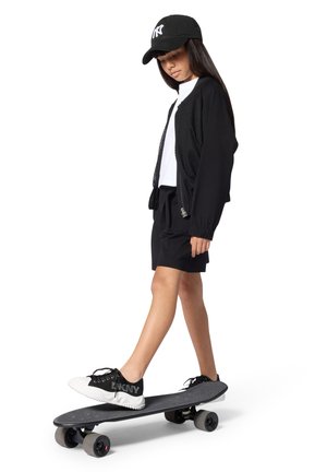 Zwarte mesh jas met ritssluiting over een wit T-shirt, gecombineerd met zwarte shorts. Zwart-witte sneakers op een grijs skateboard met zachte wielen.