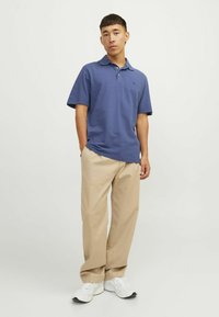 Mladic stoji s rukama u džepovima, nosi plavu polo majicu, bež hlače i bijele tenisice na jednobojnoj svijetloj pozadini.