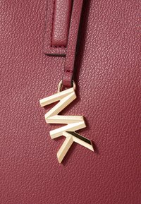 Bordeaux tekstureret lædertaske med en guldmetallisk charms med vinklede bogstaver "MK". Designet betoner et slankt, moderne æstetik.