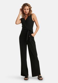 Tuta nera a coste con design a incrocio, senza maniche e vita legata. Il taglio a gamba larga conferisce una silhouette rilassata.