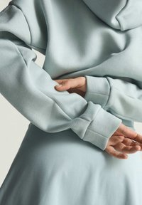 Svietlo modrá mikina s kapucňou, ktorá má jemnú textúru, rebrované manžety a voľný strih. Látka má jemný lesk a hladký povrch.