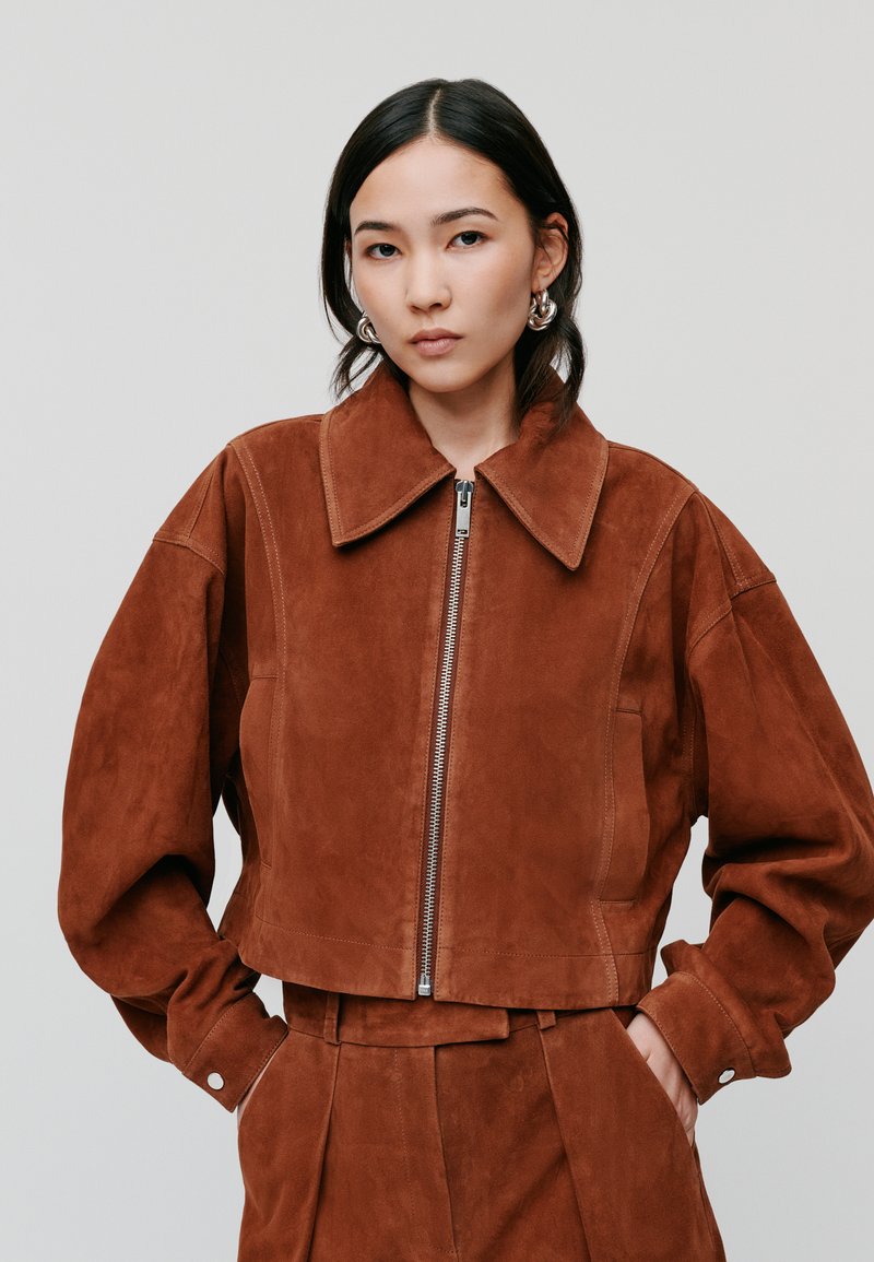 Braune Wildleder-Cropped-Jacke mit großem Kragen und Reißverschluss. Verfügt über weite Ärmel und eine glatte Textur. Minimalistisches Design.