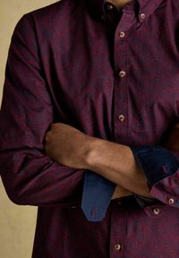 Persona ar savām rokām pāri krūtīm, tērpusies tumši sarkanā rakstainā krekla ar redzamām zilām iekšējām piedurknēm.