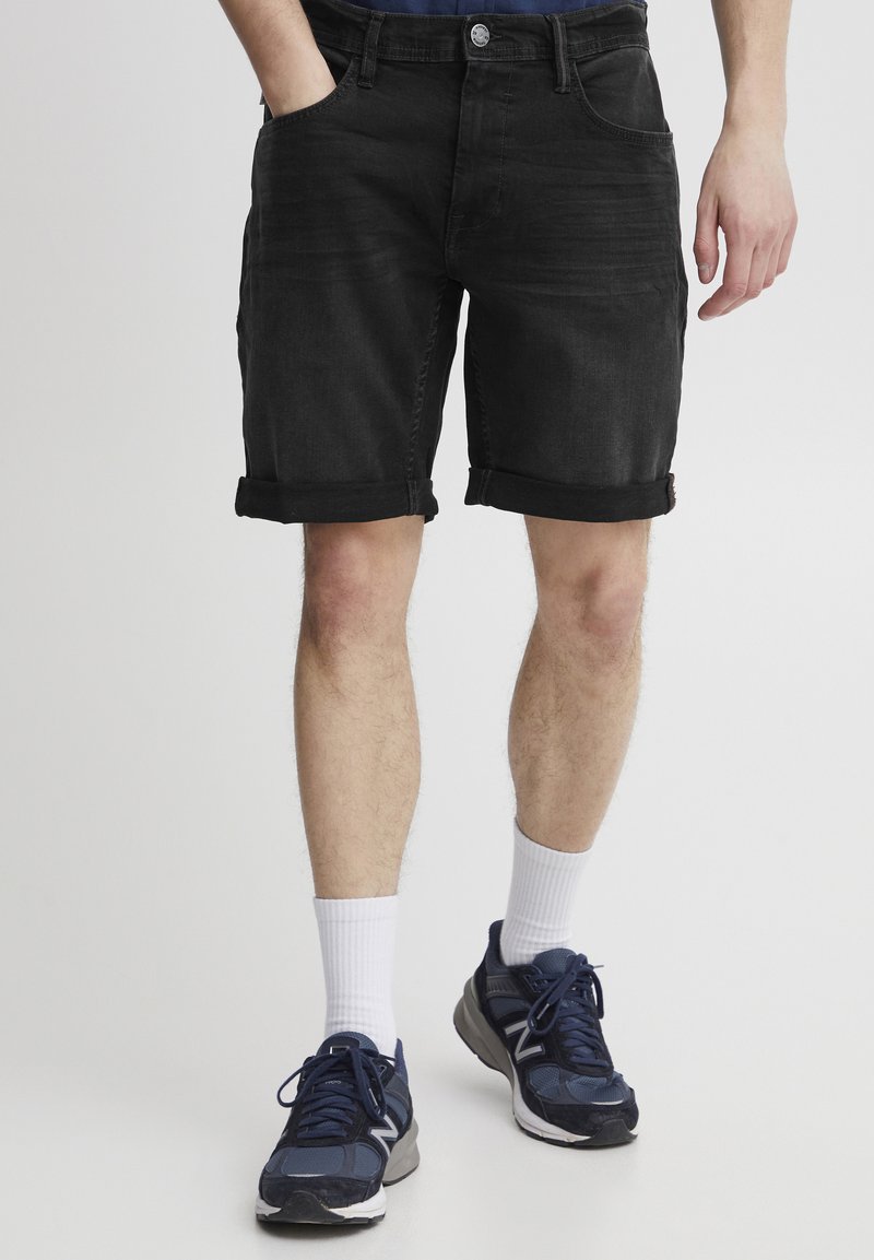 Shorts negri din denim cu tivuri rulate, având buzunare frontale și fermoar cu un buton metalic. Stilizați cu pantofi sport negri și ciorapi albi.