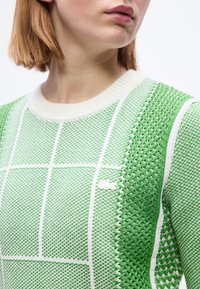 Femme portant un pull en maille à motifs verts et blancs avec un col rond, montrant le haut de la poitrine et une partie du visage tourné légèrement vers la droite.