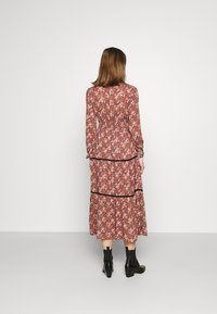 Langärmlisses Blumenkleid mit tailliertem Schnitt, gestuftem Rock, dunklem Saum und einem rötlich-braunen Hintergrund. Das Model trägt schwarze Knöchelstiefel.