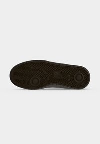 Semelle de chaussure en caoutchouc foncé avec motif circulaire et bande de roulement en forme de chevrons pour l’adhérence, photographiée sur un fond clair uni.