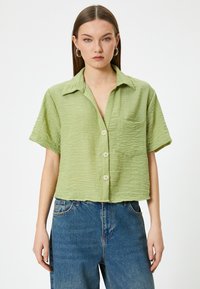 Camisa de botones de manga corta en verde claro con una tela texturizada, bolsillo en el pecho y botones de color natural. Combina con jeans rectos azules.