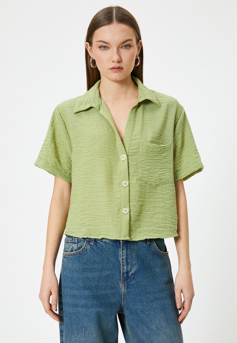 Camisa de botones de manga corta en verde claro con una tela texturizada, bolsillo en el pecho y botones de color natural. Combina con jeans rectos azules.