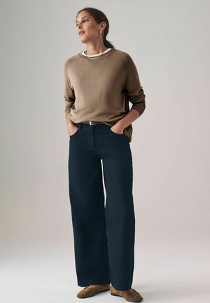 Khaki trui met een losse pasvorm en geribde manchetten, gecombineerd met brede, donkerblauwe broeken en bruine flats. Eenvoudig, textuurontwerp.