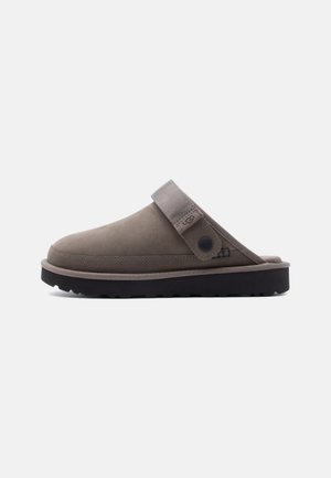 Bruine instapklomp met zwarte zool, verstelbare band met knoop aan de zijkant en een afgeronde neus.