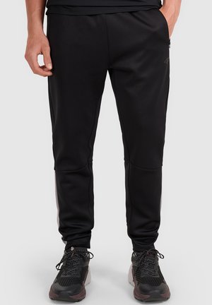 Parte inferior del cuerpo de una persona que lleva pantalones deportivos negros ajustados en los tobillos y zapatillas negras con cordones, de pie sobre un fondo liso.