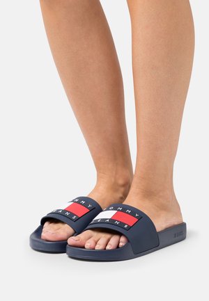 Sandalias de slide azul marino con una amplia correa decorada con acentos del logo en rojo, blanco y negro. Suela texturizada con un acabado suave.