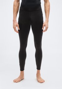 Πόδια και κορμός ενός ατόμου που φοράει εφαρμοστά μαύρα αθλητικά κολάν, όρθιο ξυπόλητο μπροστά σε απλό ανοιχτό φόντο.