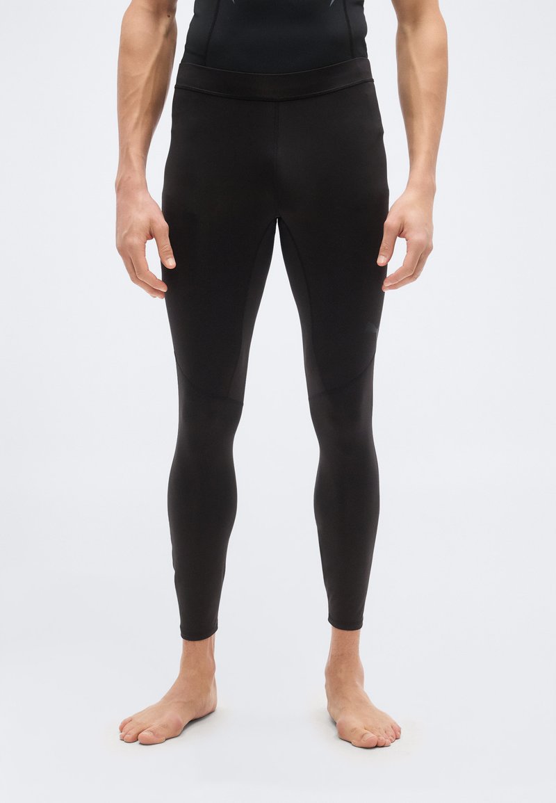 Πόδια και κορμός ενός ατόμου που φοράει εφαρμοστά μαύρα αθλητικά κολάν, όρθιο ξυπόλητο μπροστά σε απλό ανοιχτό φόντο.