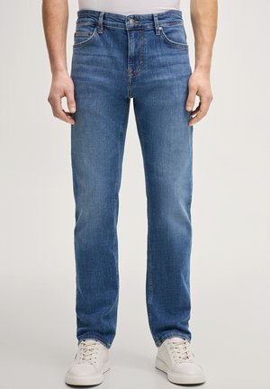 Mężczyzna ubrany w niebieskie jeansy o prostych nogawkach i białe trampki, stojący na jednolitym tle.