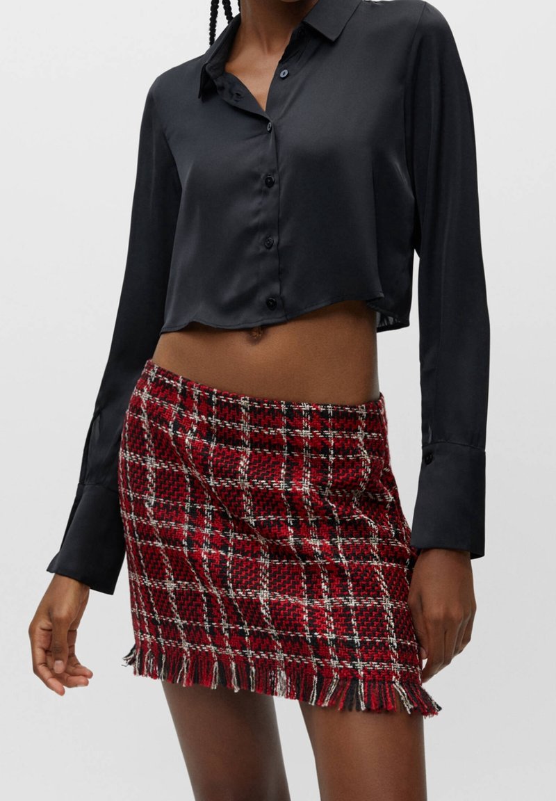 Chemisier court en satin noir avec col et fermeture à boutons, assorti à une mini-jupe écossaise rouge et noire à bords effilochés.