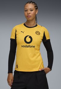 Maglia da calcio gialla con accenti neri, caratterizzata da un logo, testo dello sponsor e un delicato motivo floreale. Maniche lunghe nere sotto.