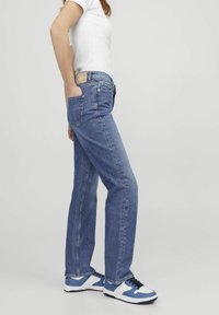 Mėlyni džinsai su tiesių kojų dizainu, turintys užpakalinius kišenes, prekės ženklą juosmens srityje ir derinami su mėlynais ir baltais sportbačiais.