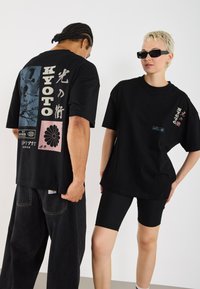 T-shirts en coton noirs surdimensionnés avec des imprimés graphiques, incluant du texte japonais et des illustrations, associés à des shorts noirs et des lunettes de soleil.