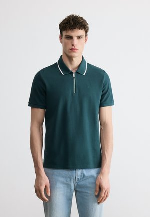 Jonge man die een donkergroene polo met korte mouwen draagt, met witte rand aan de kraag en lichtblauwe spijkerbroek, staand tegen een effen achtergrond.