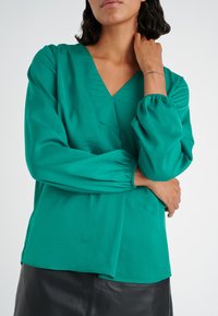 Donna che indossa una blusa verde teal a maniche lunghe con scollatura a V e polsini elasticizzati, in piedi con le braccia incrociate e una mano che tocca il collo contro uno sfondo neutro.