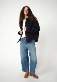 Marineblaue Wolljacke mit Knopfverschluss, entspannter Schnitt, getragen über einem weißen Hemd und hellblauen weit geschnittenen Jeans, kombiniert mit braunen strukturierten Schuhen.