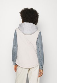 Quiltet vest med en beige krop, denimærmer og en grå hætte. Har knapper på ærmerne og en afslappet silhuet.