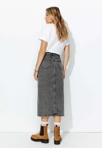 Femme aux cheveux longs portant un t-shirt blanc, une jupe midi en jean gris taille haute, des chaussettes blanches et des bottines brunes, se tenant de dos.