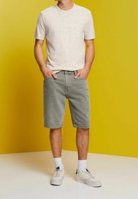 Chemise à manches courtes tricotée texturée de couleur claire associée à un short en denim gris. Le modèle porte des baskets beige avec des chaussettes blanches sur un fond jaune.