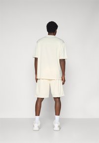 Cremefarvet træningssæt bestående af en løs kortærmet top og shorts til knæene, lavet af blødt, glat stof, vist bagfra.