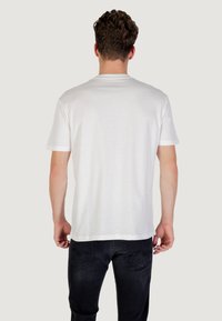 Camiseta de algodón blanca con cuello redondo y mangas cortas, mostrada desde atrás, combinada con jeans oscuros. Textura suave y diseño sencillo.