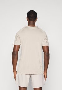 Ανοιχτό μπεζ αθλητικό μπλουζάκι με ανάγλυφες δικτυωτές πλάκες στους ώμους, κοντά μανίκια και στρογγυλή λαιμόκοψη, συνδυασμένο με ανοιχτόχρωμες βερμούδες.