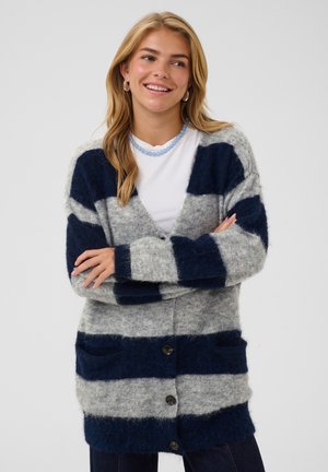 Jeune femme souriante, les bras croisés, portant un cardigan duveteux gris et bleu marine rayé sur une chemise blanche et un collier de perles bleu.