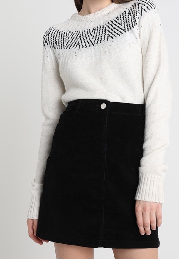 Femme portant un pull en maille blanc avec des motifs géométriques noirs et une jupe noire taille haute avec un bouton à l'avant.