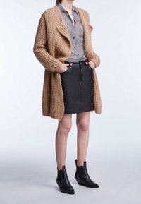 Cardigan beige tricoté, chemise à carreaux noire, jupe en cuir noire et bottines noires. Tissu texturé et design en couches.