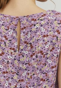 Lilla blomstermønstret stof med små blomster i gul, hvid og rød. Nøglehulslukning bagpå med en rund lilla knap.
