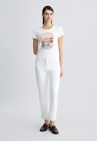 T-shirt blanc avec un imprimé graphique coloré, associé à un pantalon blanc avec un ourlet en dentelle festonnée et des chaussures bateau marron.