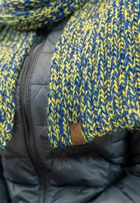 Strickschal mit blauen und gelben Fäden, strukturiertem Muster und einem braunen Lederetikett; getragen über einer schwarzen Daunenjacke.