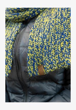 Strickschal mit blauen und gelben Fäden, strukturiertem Muster und einem braunen Lederetikett; getragen über einer schwarzen Daunenjacke.