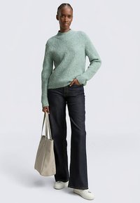 Hellgrüner, gerippter Pullover mit hohem Kragen, kombiniert mit dunklen, ausgestellten Jeans. Das Modell hält eine beige Tragetasche und trägt weiße Turnschuhe.
