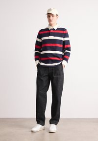 Stripet langærmet polo-shirt i navy, rød og hvid, med krave og frontlomme, parret med mørke denimbukser og hvide sneakers.